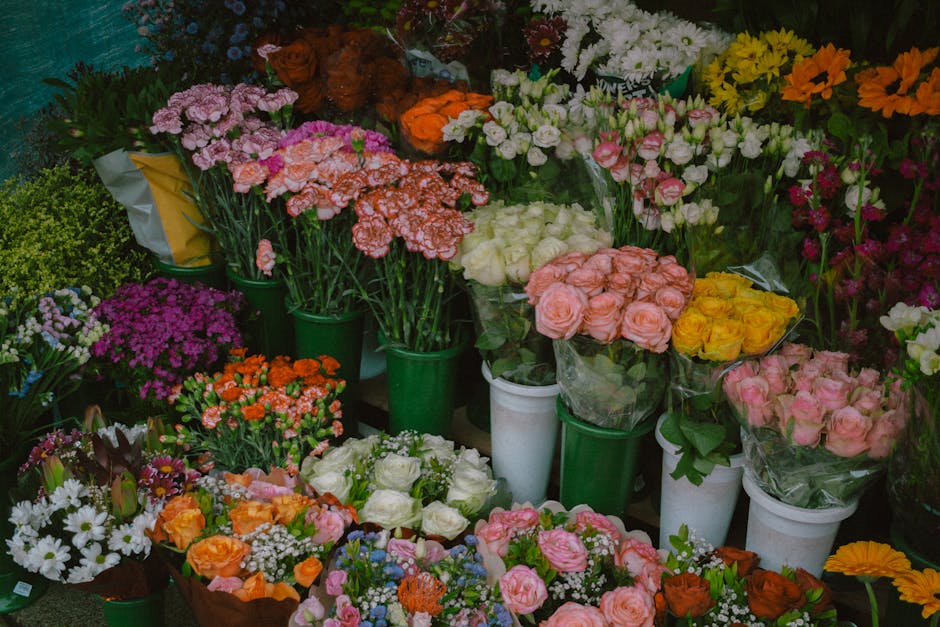A vibrant display of fresh flower bouquets arranged in green and white vases and wrapped in clear plastic, featuring roses in soft pink and yellow, carnations in coral and white, daisies, and other se