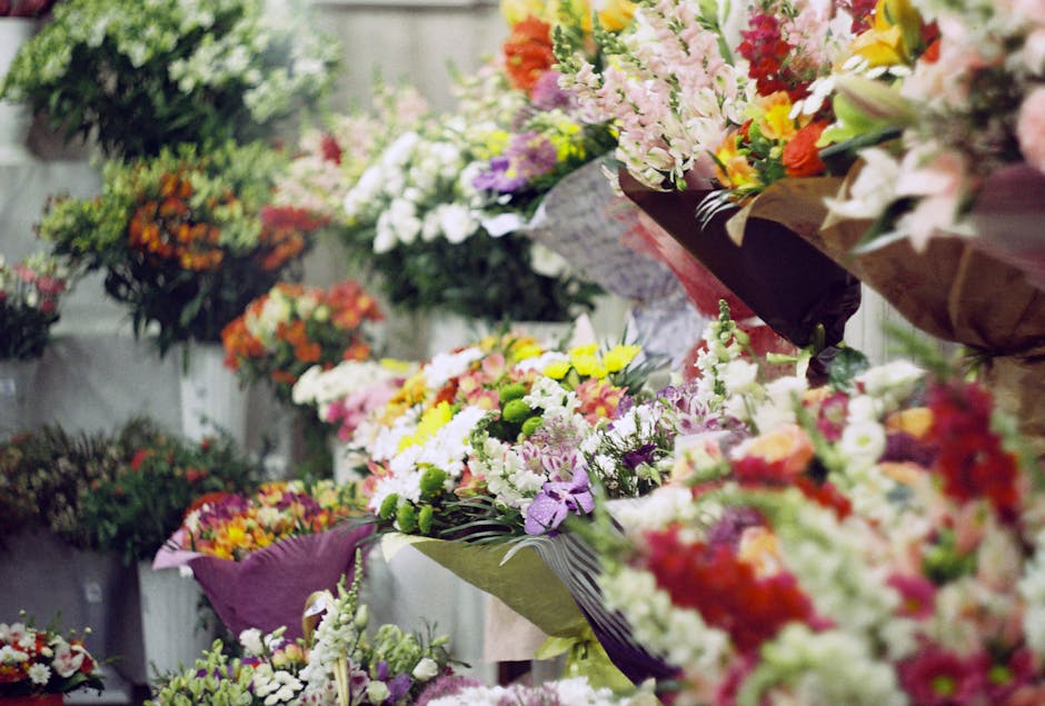 A vibrant display of fresh floral bouquets arranged at a flower shop, featuring a variety of flowers including white, pink, purple, red, orange, and yellow blooms. The bouquets are wrapped in colorful
