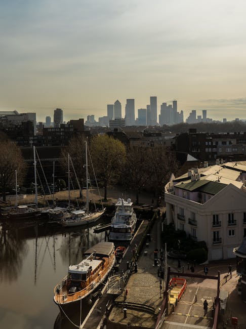 This image features a cityscape view of Millwall with a canal in the foreground, alongside a marina filled with sailboats and a pier. The scene is captured during the daytime under cloudy skies, with
